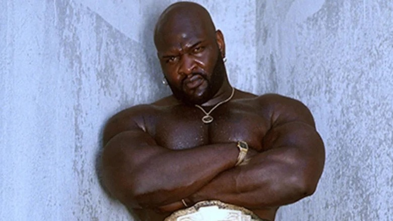 Ahmed Johnson poses in front of a wall wearing a championship belt.
