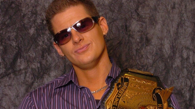 Eric Priest poses for the camera wearing sunglasses and carrying the AAW Heritage Championship belt across his shoulder.