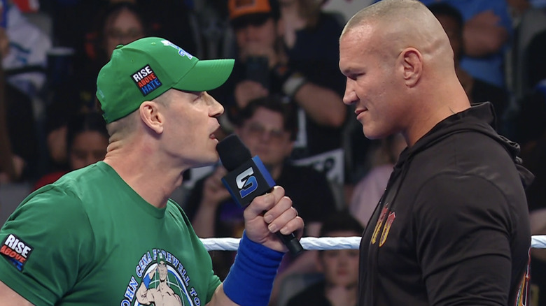 John Cena leans in to a smirking Randy Orton as he speaks on the microphone.