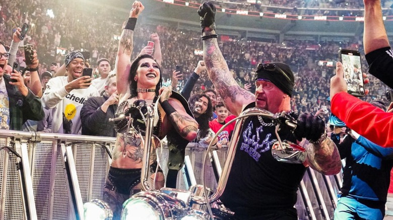 Women's World Champion Rhea Ripley stands beside The Undertaker, both of their arms raised in victory, with Undertaker riding a motorcycle, on "WWE Raw."