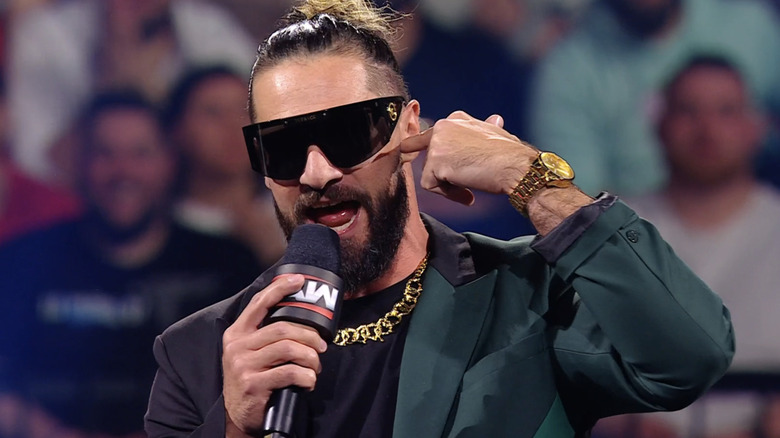 Seth Rollins speaks on the microphone in a black and dark green suit, gold jewelry, and large black sunglasses.