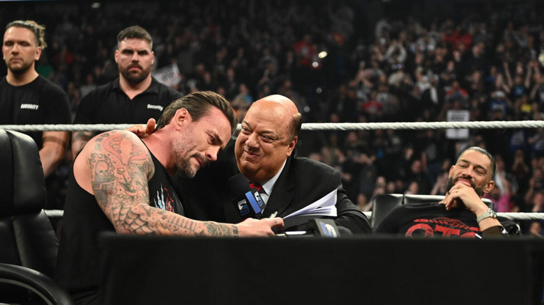 CM Punk is held close by a smiling Paul Heyman as Roman Reigns watches in the background of a contract signing.